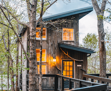 Wooden two-story treehouse lodging in the evening with light from the interior lights giving a soft glow through the windows.